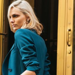 Volumising spray: Woman with volumised blonde medium-length hair, wearing teal blue suit jacket while posing outside a golden hotel