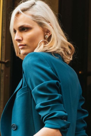 Volumising spray: Woman with volumised blonde medium-length hair, wearing teal blue suit jacket while posing outside a golden hotel