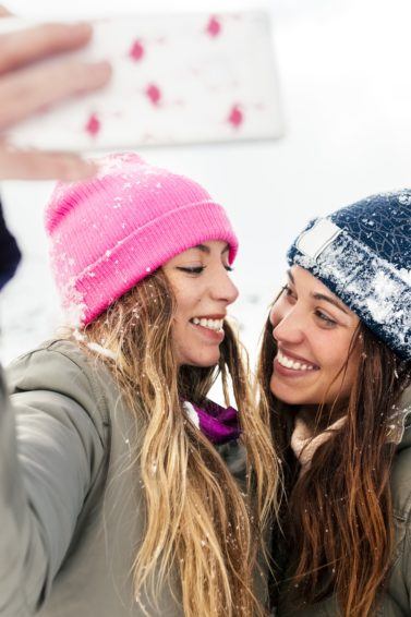close up shot two women in the snow with jackets, with wavy hair and winter hats taking a selfie