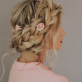 Woman with long golden blonde hair styled into a milkmaid braid updo with pink roses in it, wearing a pink dress and posing against a grey wall