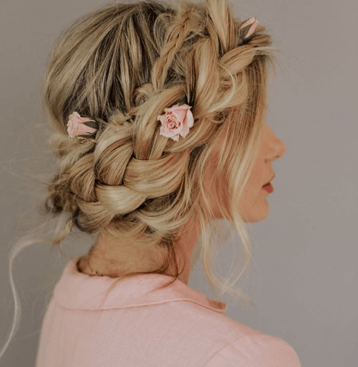 Woman with long golden blonde hair styled into a milkmaid braid updo with pink roses in it, wearing a pink dress and posing against a grey wall