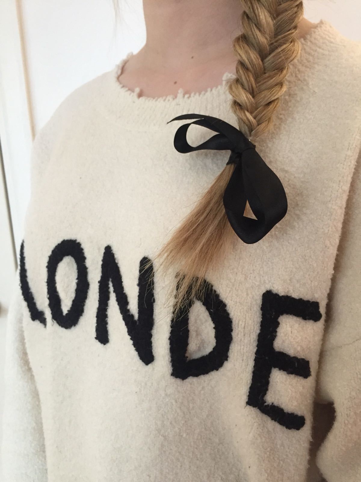 close up shot of woman with blonde hair styled into a side fishtail braid with a bow accessory on it, wearing a jumper that says blonde on it
