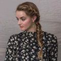 Braided updos for long hair: Woman with long dark blonde hair styled into a side boho braid updo, wearing floral top and posing against a grey wall in a studio
