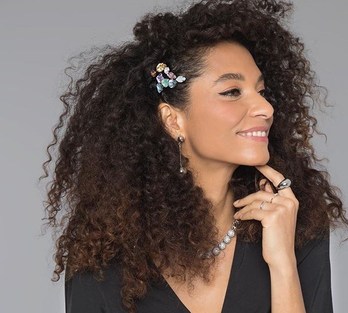 Woman with long natural curls styled with a hair accessory, wearing all black and posing in the studio