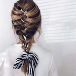 Woman with long bronde hair styled into a French braid updo with a scarf weaved through it, wearing white jumper and posing against a white wall