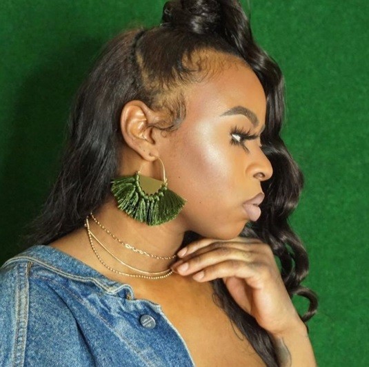 woman with her dark brown hair in a half up curly ponytail wearing a denim jacket and statement green earrings