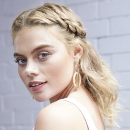 New Year's Eve hair ideas: Blonde model with wavy mid length hair in a twisted half-up half-down style, wearing a white cami top and standing against a white brick wall