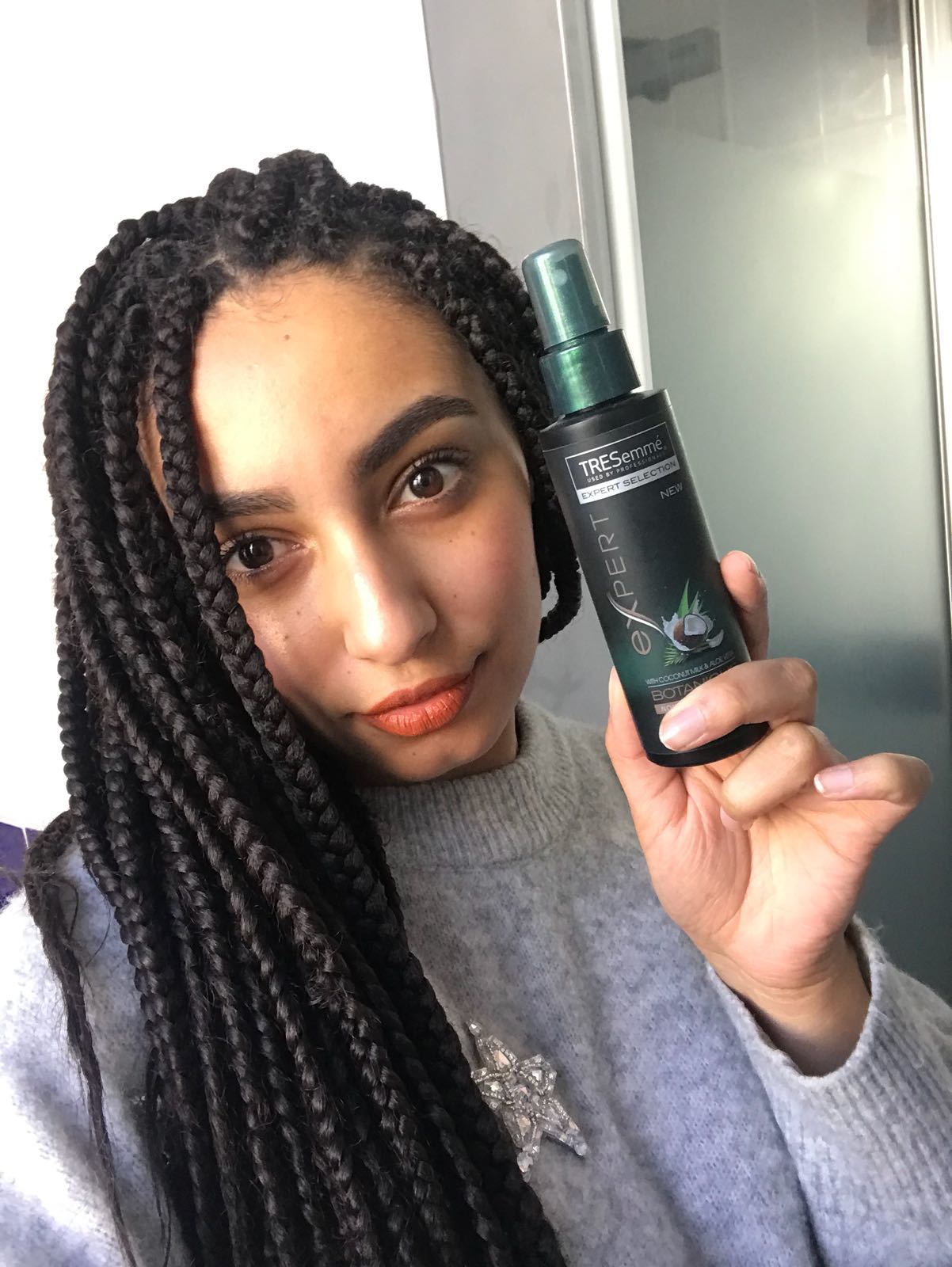 All Things Hair editor Amra with long box braids, swept to the side and wearinng silver jumper holding a tresemme leave in conditoner