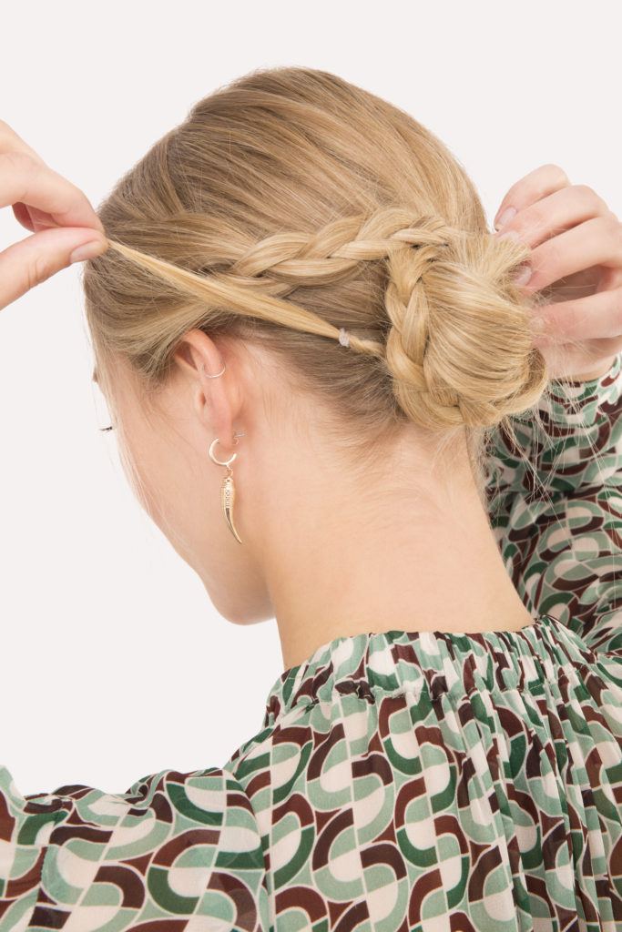 Braided updo blonde girl pinning plait around bun with back to camera