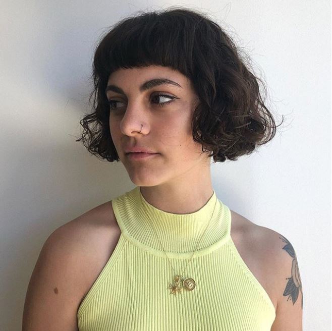 2019 haircut predictions: Woman with a curly blunt short bob with micro bangs wearing a yellow vest.