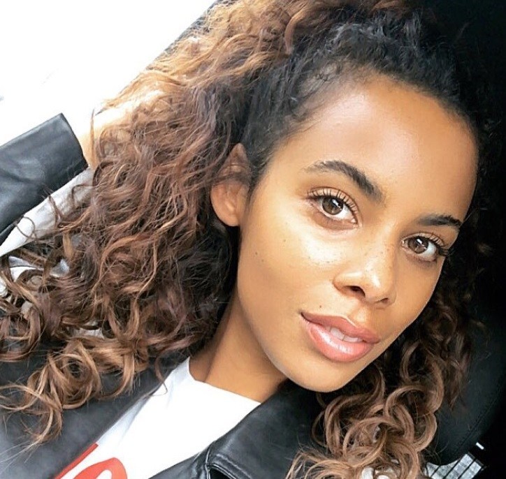 close up shot of rochelle humes with natural curly hair styled into a half up half down ponytail, wearing black jacket and posing