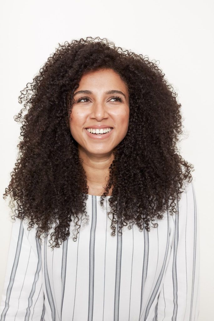 woman with long dark brown curly hair wearing a striped outfit smiling and looking in the distance