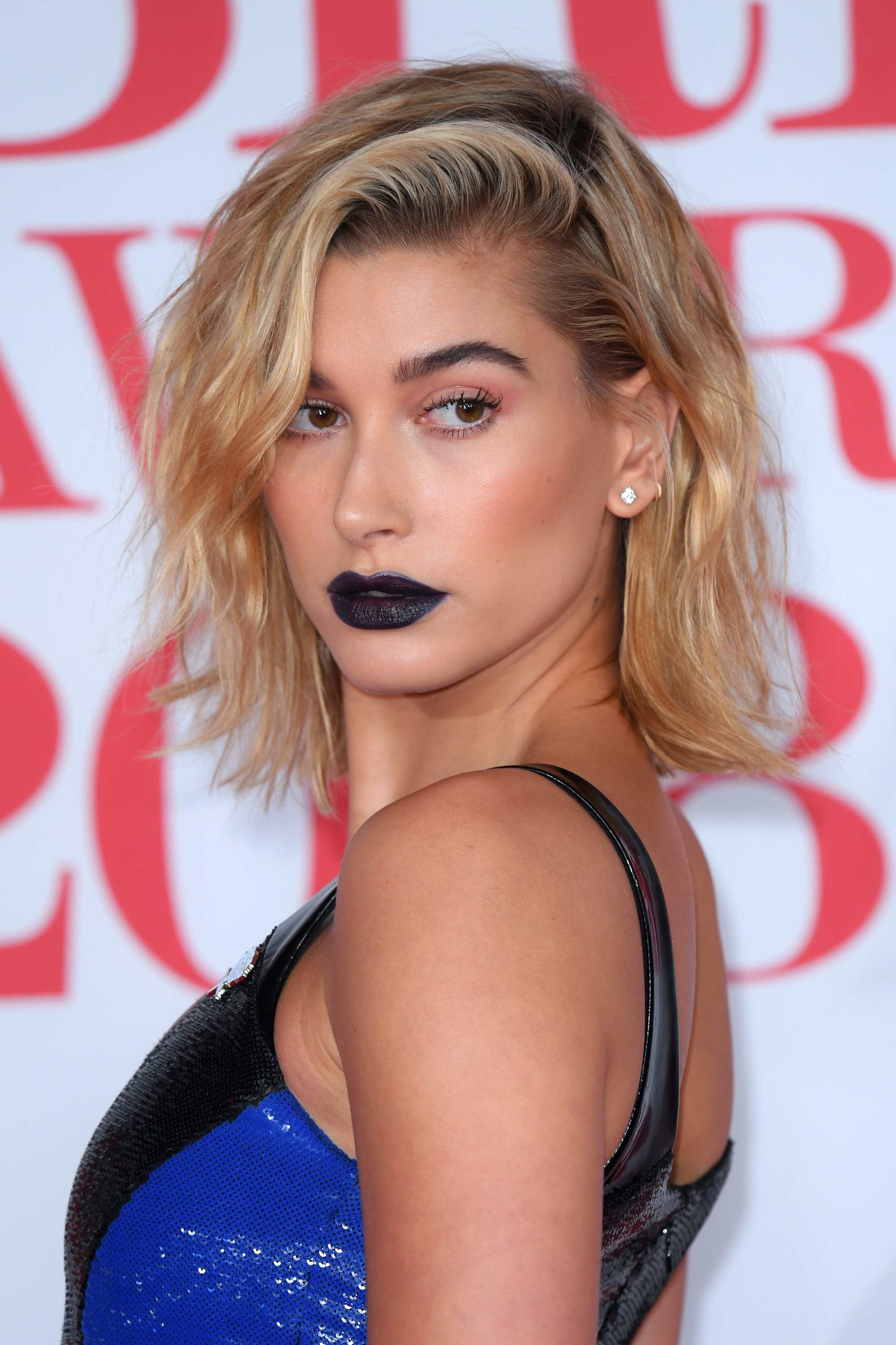 close up shot of hailey baldwin with beachy short waves and wearing blue and black dress with dark blue lipstick, posing on the brit awards 2018 red carpet
