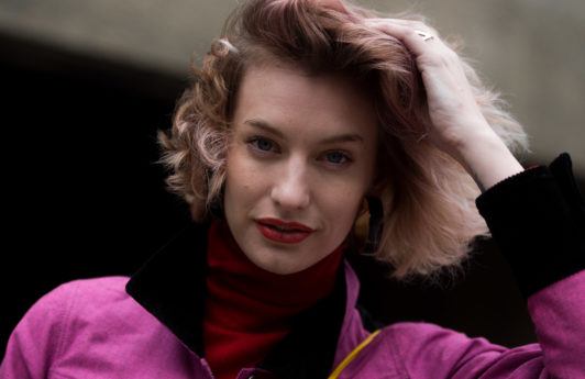 best hair mousse guide: close up street style shot of woman with pastel pink wavy bob, wearing pink jacket and red scarf