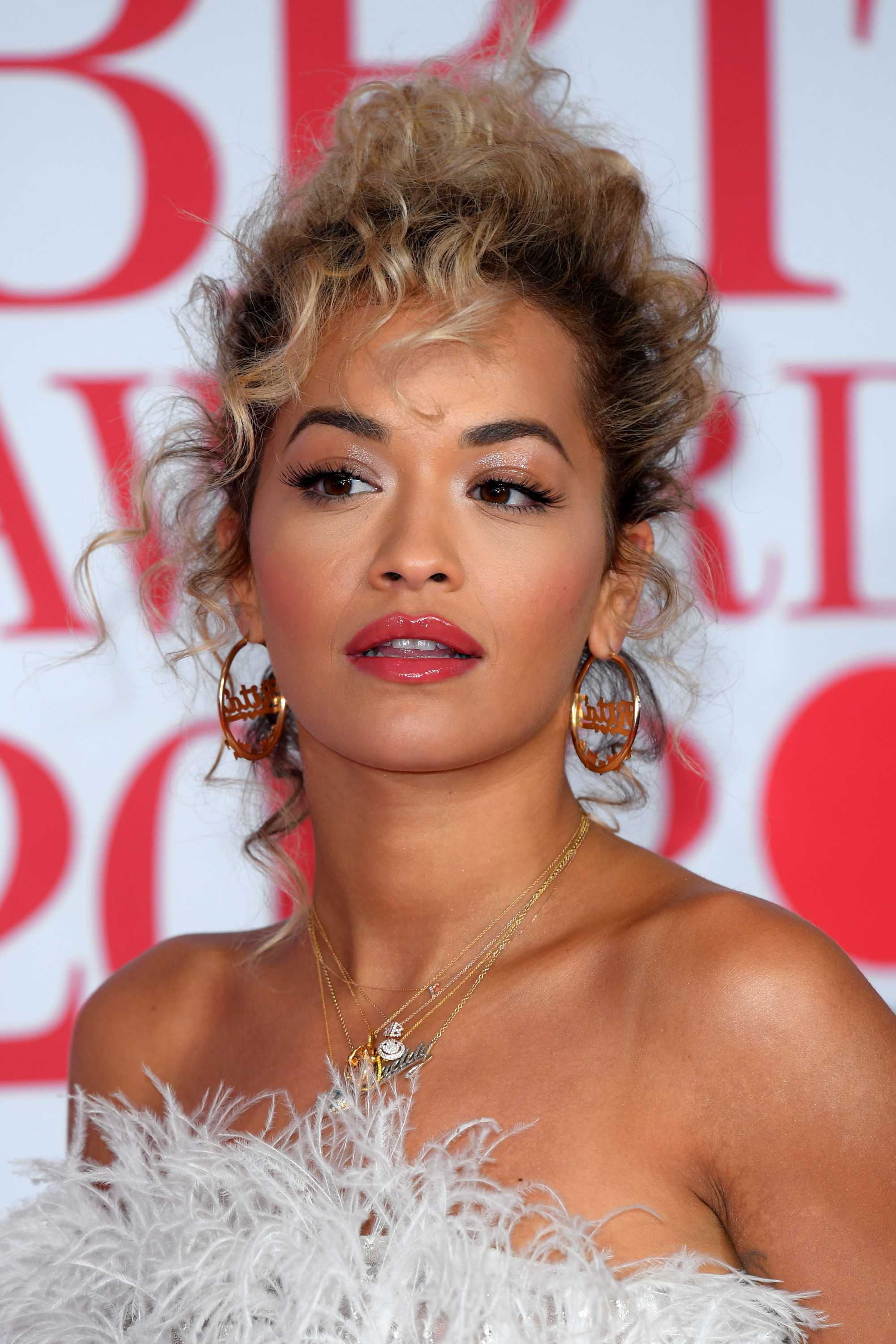 close up shot of rita ora with curly updo hairstyle, wearing hoop earrings and white frilly dress on the brit awards 2018 red carpet