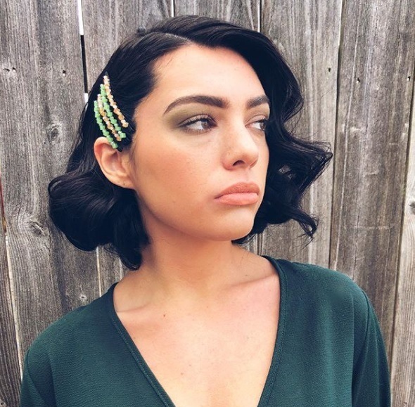 Short night out hairstyles: Woman with short dark brown hair styled into vintage curls, wearing colourful bobby pins in her hair with a green wrap dress, posing outside