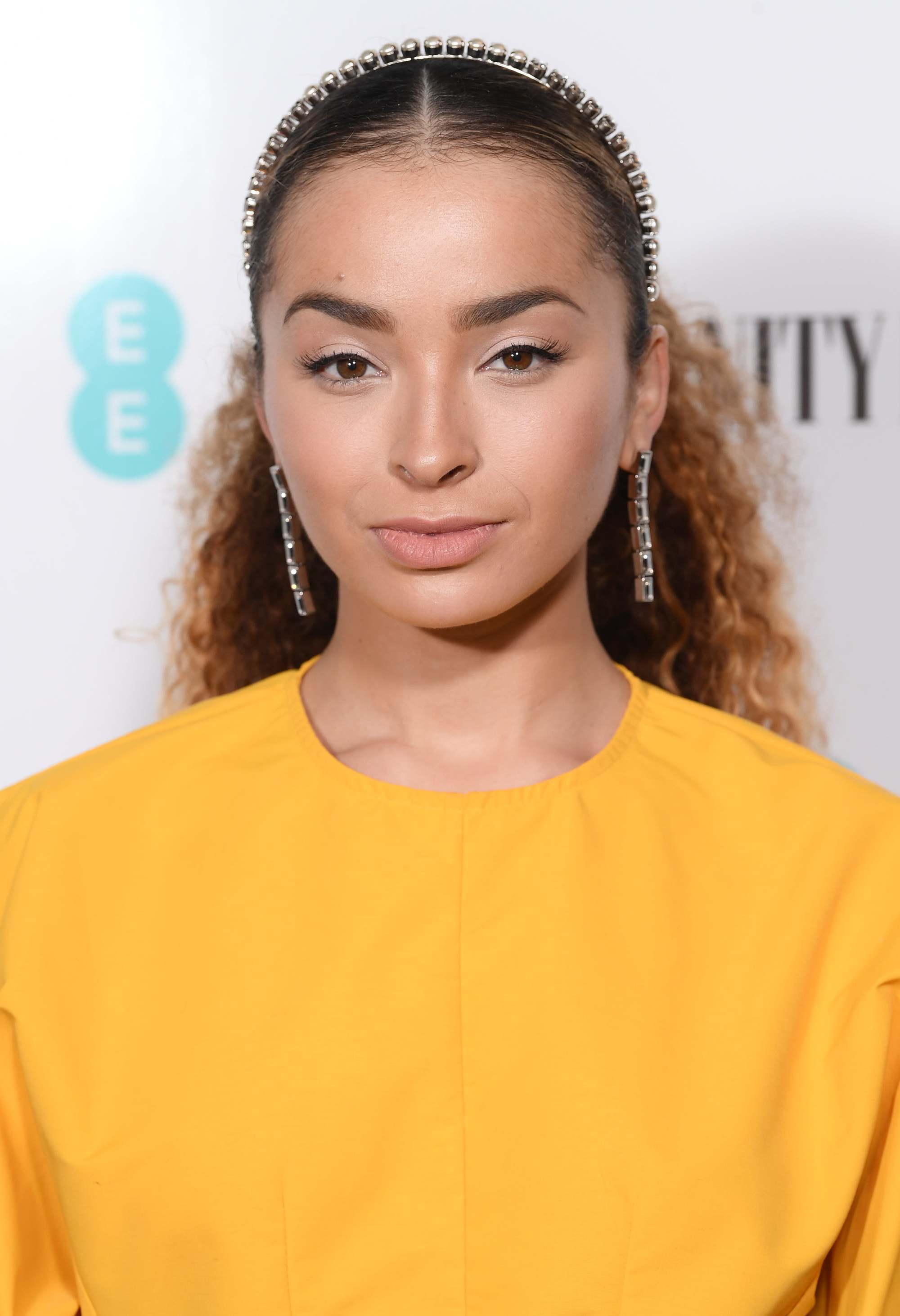 Celebrity Valentine's Day Hairstyles: Shot of Ella Eyre with long curly blonde hair styled into a sleek ponytail, wearing a headband with a yellow dress on the red carpet