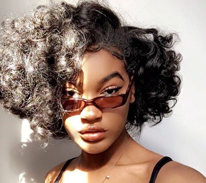 close up shot of woman with short curly weave hairstyle, wearing micro sunglasses and posing in the sunlight