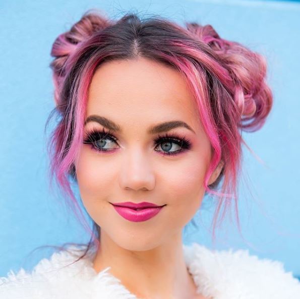 woman with pink hair in high space buns wearing white fluffy jacket against baby blue wall