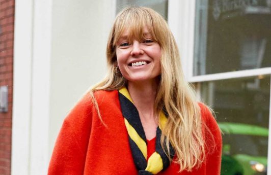 Spring haircuts: Blonde street styler with mid length wavy hair and a full fringe, wearing an orange red jumper and a striped neck scarf, standing outside in the street