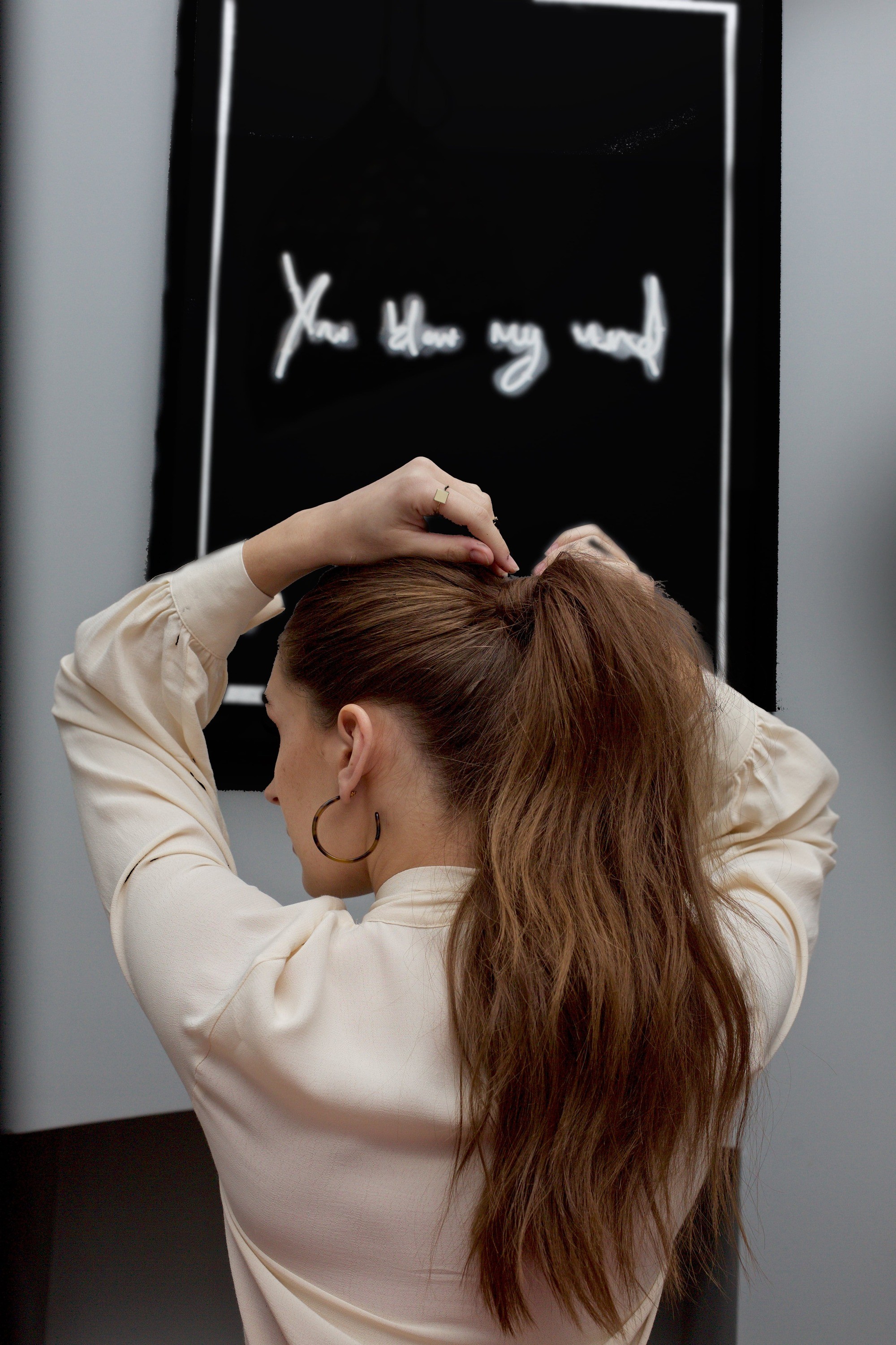 Spring haircuts: Back view of a brunette woman with a long voluminous wavy ponytail, putting a bobby pin in her hair and wearing a cream blouse