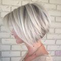 stacked bob haircuts: close up shot of woman with stacked silver blonde hair, posing and wearing brown jumper