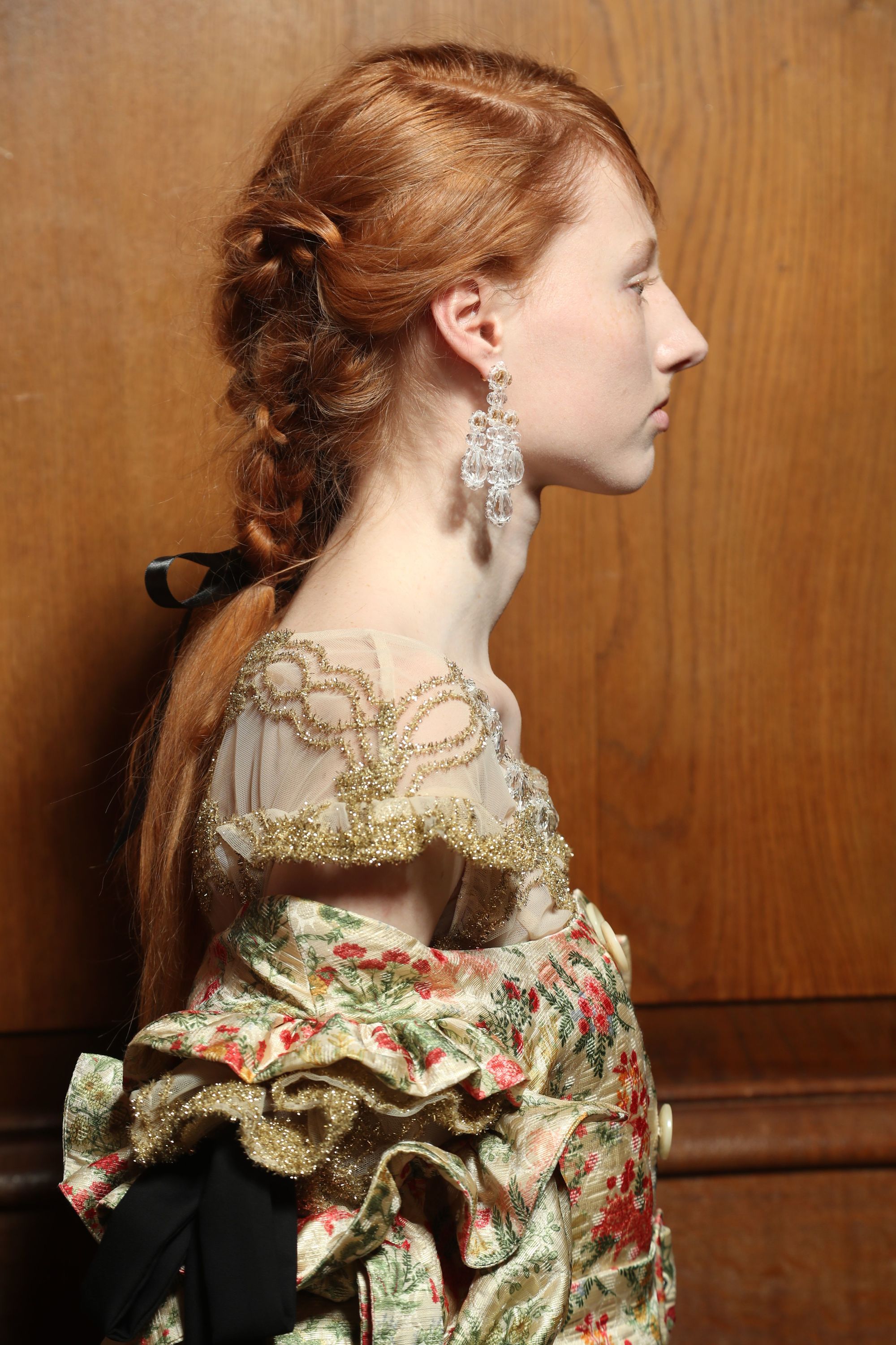 close up shot of model backstage with braided ponytail, complete with a hair bow accessory, wearing floral gown and drop earrings