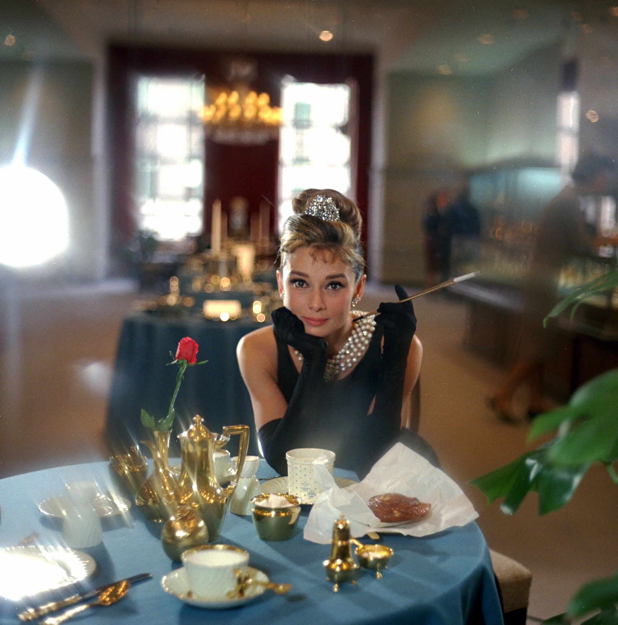 Audrey Hepburn as Holly Golightly, with beehive updo hairstyle, wearing all black with pearls and posing in a cafe