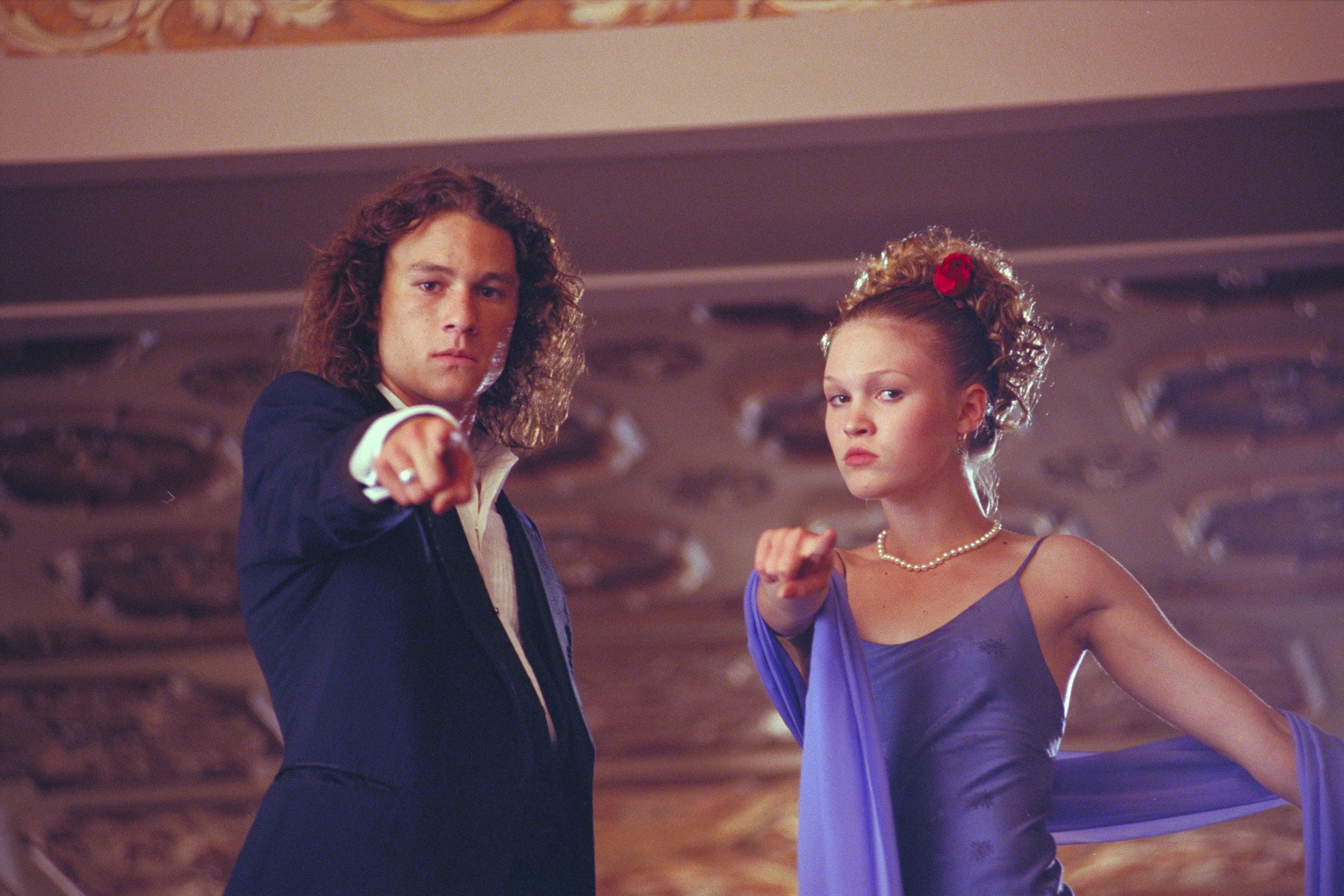 close up shot of julia stiles with curly updo hairstyle with flowers in it, wearing all purple, standing next to heath ledger on the set of 10 Things I hate about you