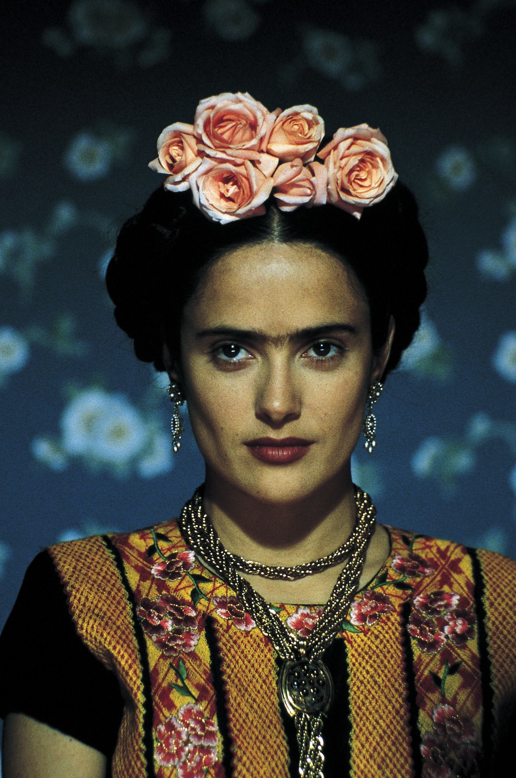 close up shot of selma hayek as frida kahlo, with milkmaid braid updo with flowers in it, wearing drop earrings and floral outfit