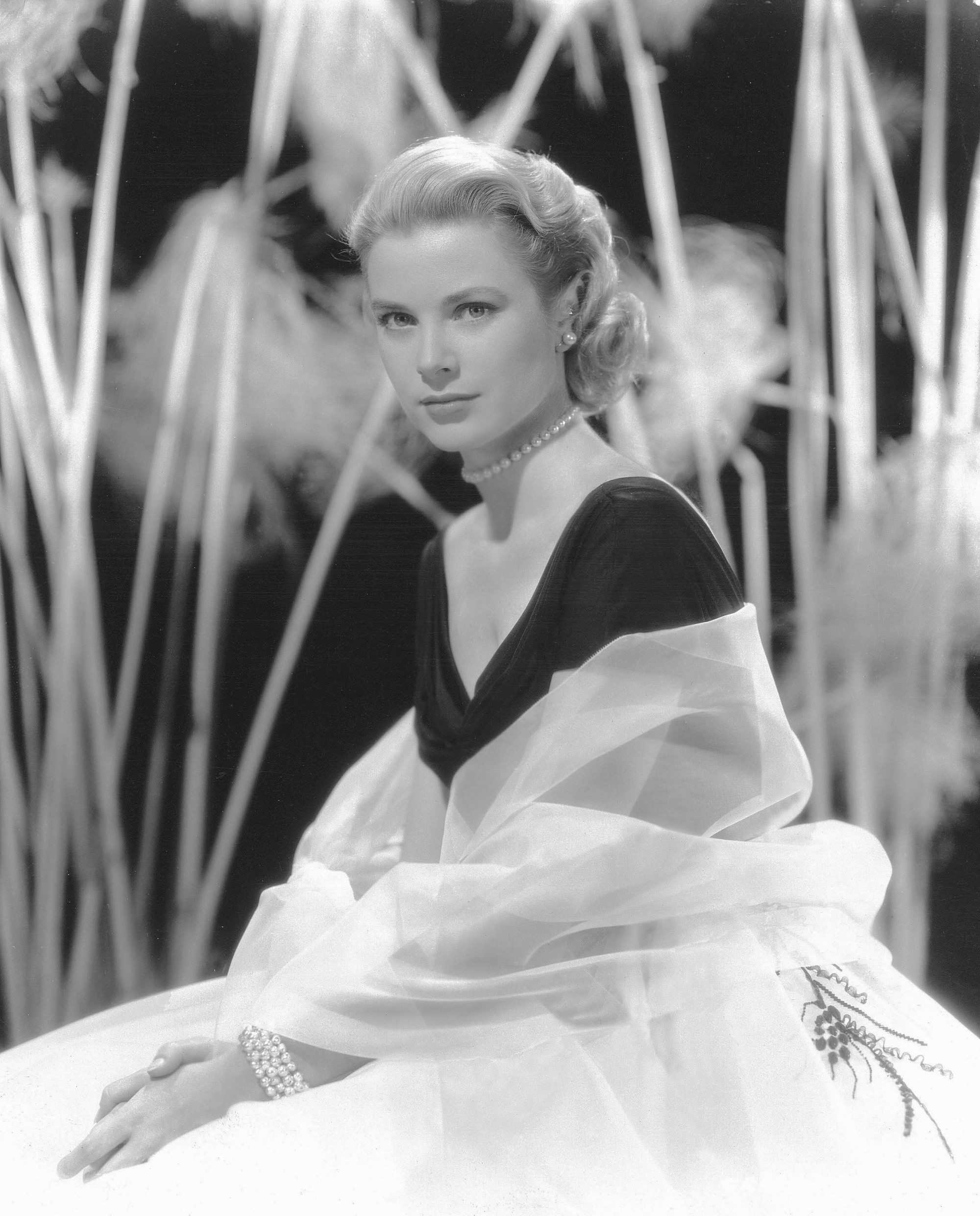 Grace Kelly coiffed blonde shoulder length hair in smooth back retro waves wearing black v-neck top with white sheer wrap