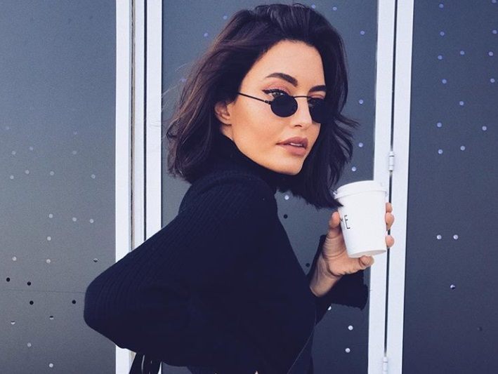 close up shot of woman with short textured dark brown hair, wearing all black, micro sunglasses and holding a cup