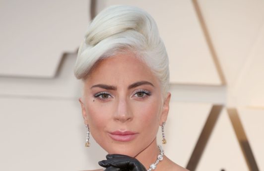 Oscars 2019 hairstyles: Lady Gaga at the 2019 Oscars with her platinum blonde hair in an Audrey Hepburn inspired chignon updo, wearing a black strapless dress and long black gloves