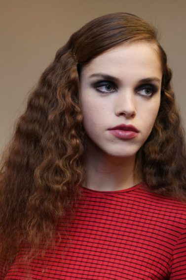 models backstage with medium length hair in side parting with smooth roots and wavy lengths wearing red jumper and denim jacket