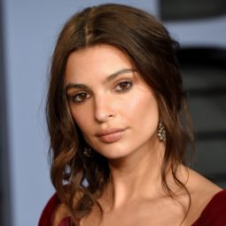 Side braid hairstyles: Emily Ratajkowski with brown wavy hair in a loose side braid with burgundy ribbon at the end.