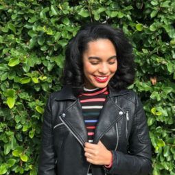 girl with relaxed and curled natural hair wearing a leather jacket and stripy rainbow roll neck standing in front of a hedge