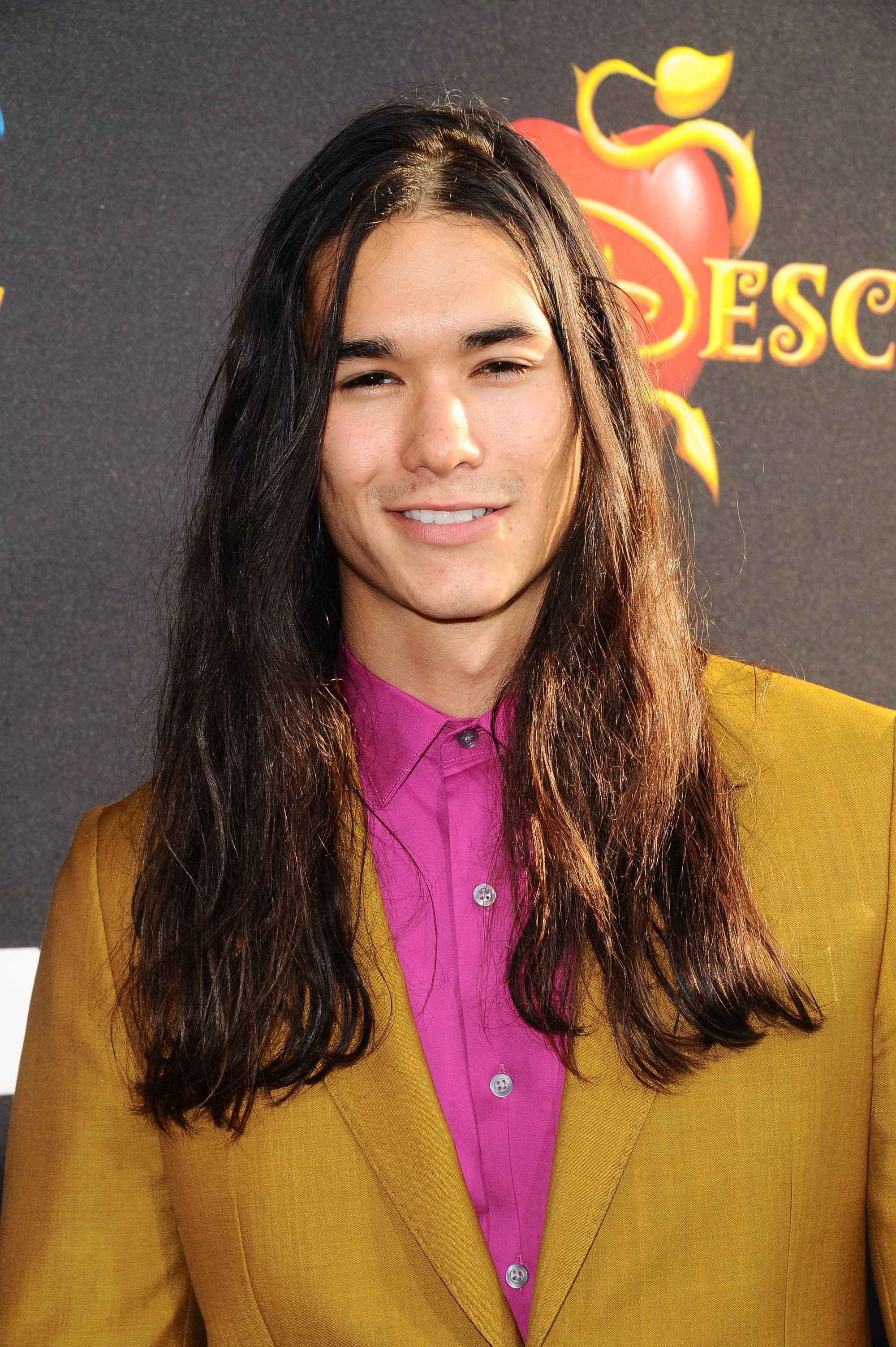 twilight actor booboo stewart on the red carpet with his long wavy dark hair wearing a pink shirt and mustard blazer