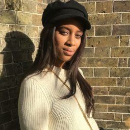 close up shot of woman with long relaxed wavy hair wearing a baker boy hat, white jumper and posing outside