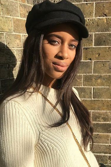 close up shot of woman with long relaxed wavy hair wearing a baker boy hat, white jumper and posing outside