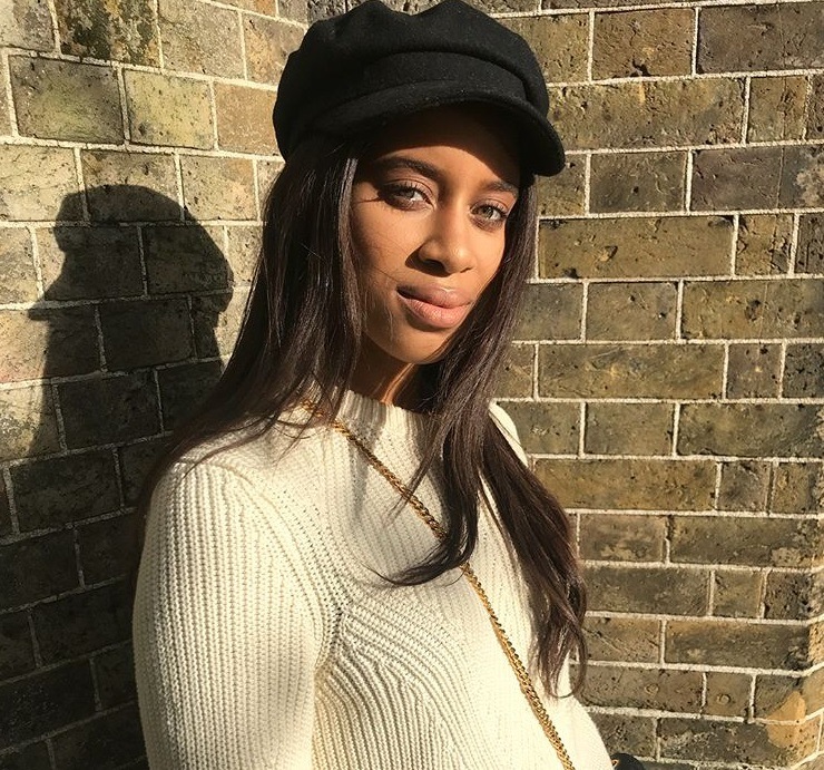 close up shot of woman with long relaxed wavy hair wearing a baker boy hat, white jumper and posing outside