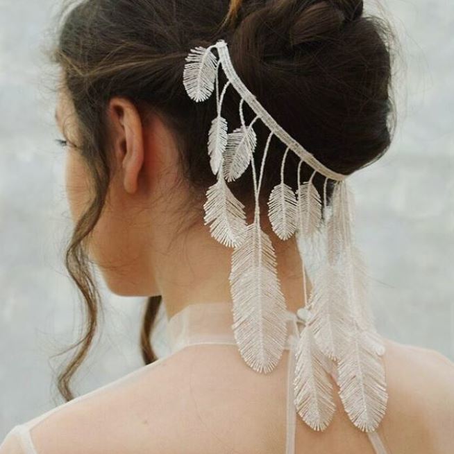 back side view of woman with dark brown hair in updo with white feather hanging detailing wearing sheer white wedding dress