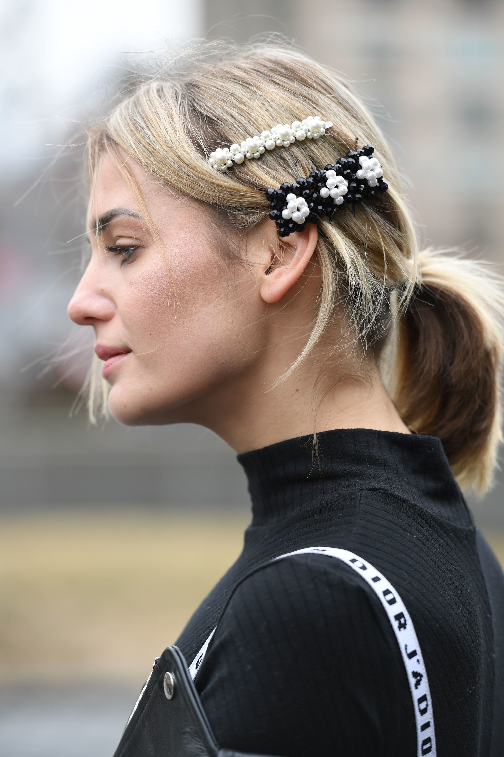 Growing out bangs: Street style woman with blonde medium length hair in low ponytail with pearl hair clips to one side wearing a black top.