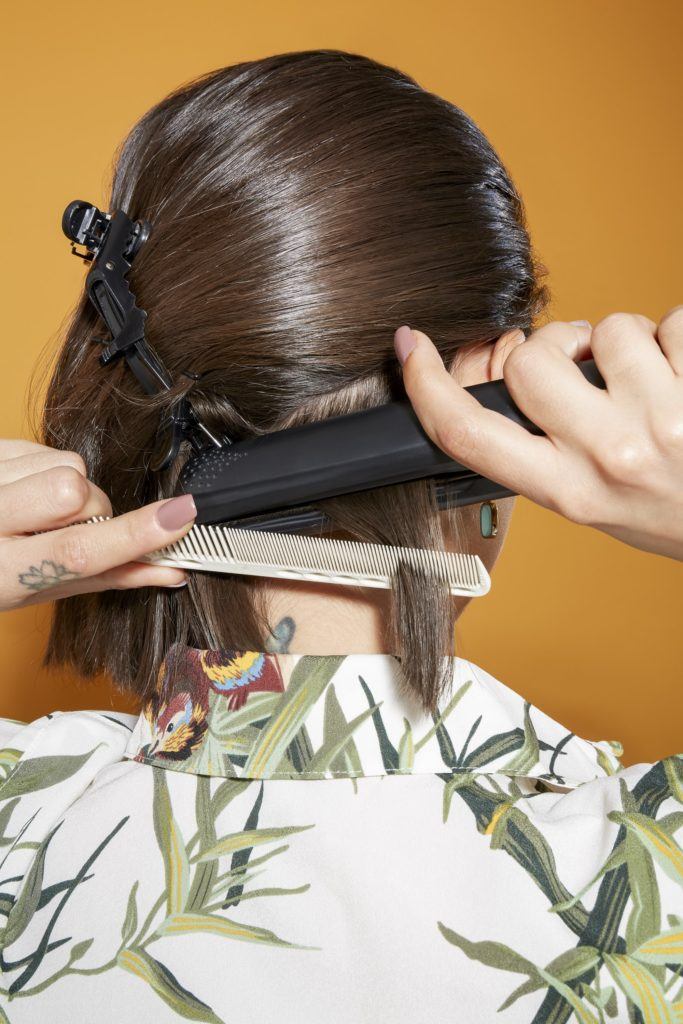 back view of a woman with shoulder length dark brown hair using a straightener and a comb to style her hair