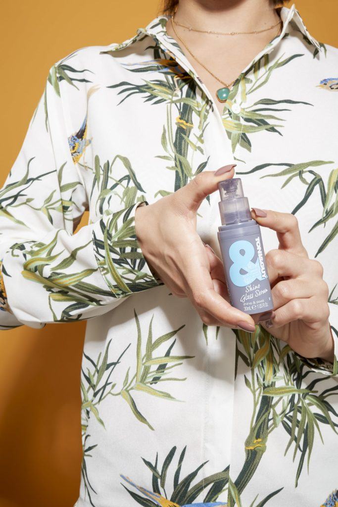 woman in a palm tree printed shirt holding a bottle of Toni & Guy shine gloss serum