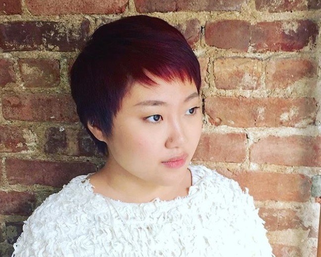close up shot of woman with feathered pixie haircut and red violet hair, wearing white top and posing against a brick background