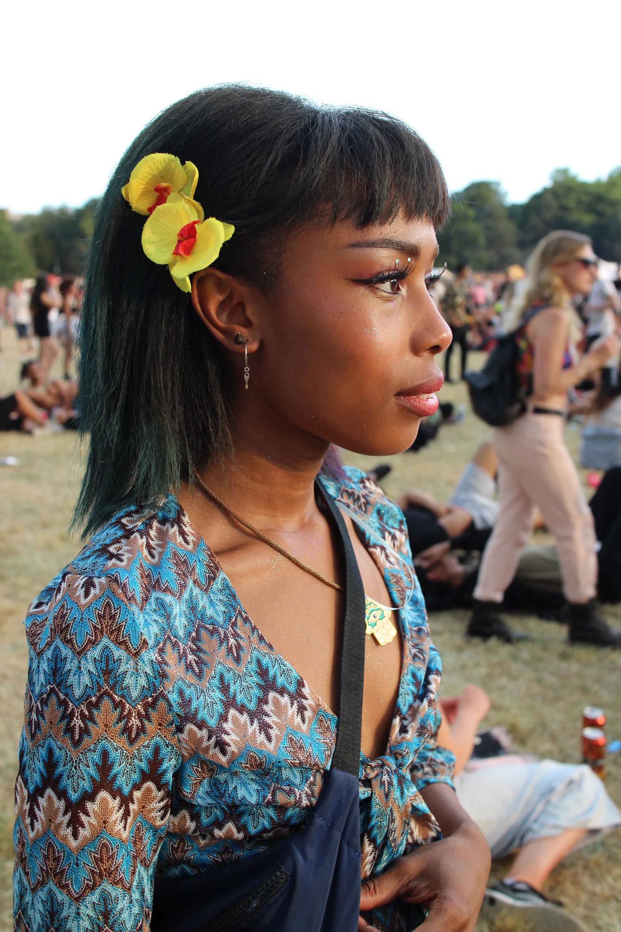 Floral festival hair: side view of black woman with straight dark brown hair at festival with yellow sunflower hairclips positioned on the side of her just behind her fringe. Girl is wearing a blue patterned top and side bag.