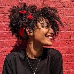 Transitioning hairstyles: Woman with curly dark brown pineapple updo, wearing black and posing against red brick wall