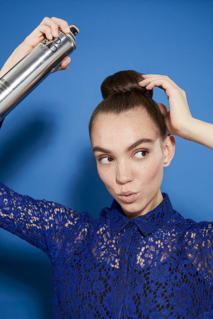 woman with brunette hair in a tight bun spraying it with hairspray, wearing a blue lace outfit