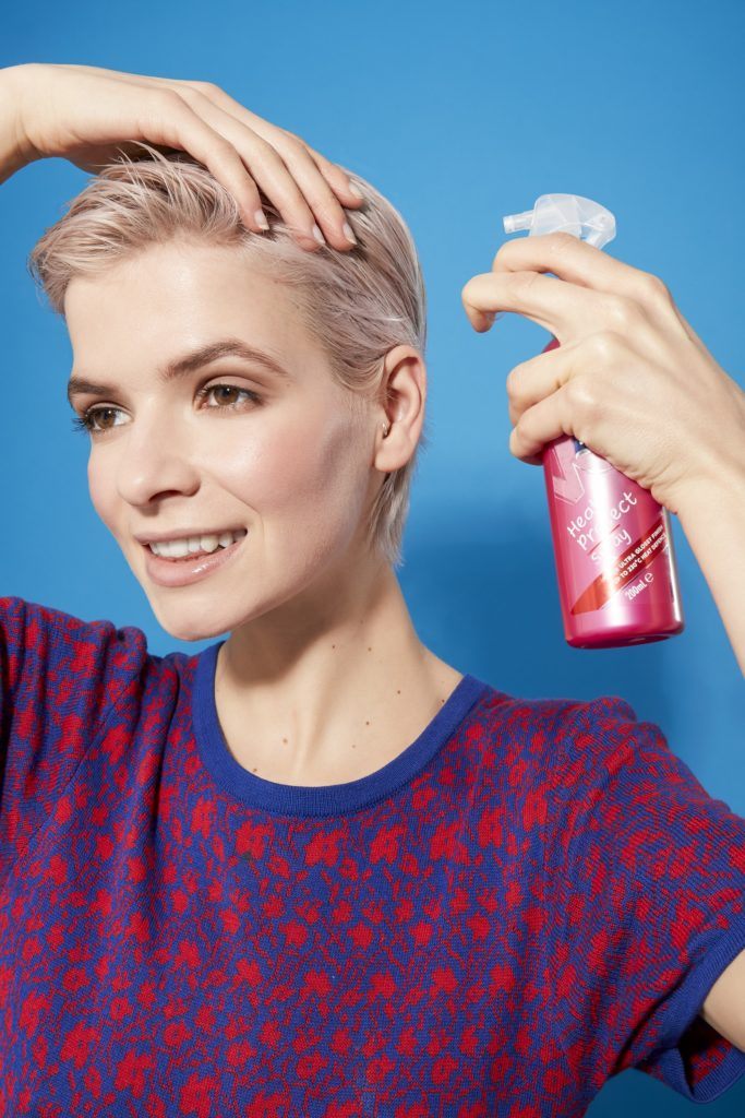 woman with platinum blonde hair in a pixie cut style, spraying a heat protectant into her hair