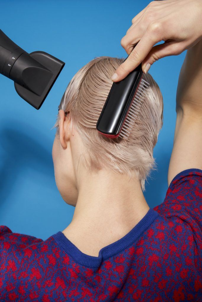 back view of a woman with platinum blonde hair in a pixie cut style using a brush and hairdryer to style her hair